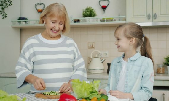 Senior grandmother is making salad and feeding cute happy child with vegetable chops expressing love having fun in kitchen at home. People and lifestyle concept.