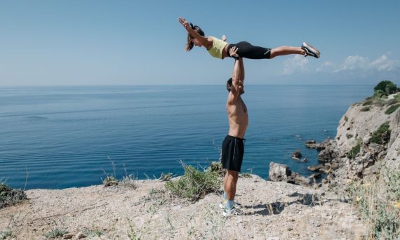 A couple performs acrobatic exercise on a scenic ocean cliff, embodying fitness and adventure.